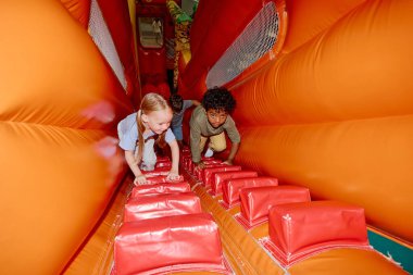 Küçük sevimli kültürler arası arkadaşlar oyun parkında kırmızı merdivenlerle yumuşak turuncu deri tünele tırmanırken dik kaymaya devam ediyorlar.