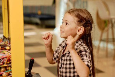 Uzun sarı saçlı neşeli küçük kız lolipoplu otomat bardağının içinden bakıyor ve bir tane vinç yapmaya çalışıyor.