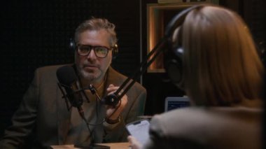 Orta yaşlı beyaz erkek politikacı ya da kulaklıklı bilim adamı stüdyoda podcastda konuşurken, kahve içerken, akıllı telefonlu kadın sunucunun sorularını cevaplarken.