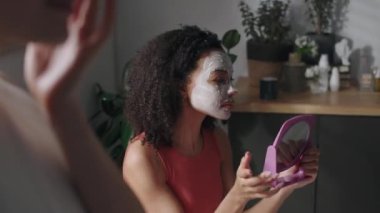 Göğüs hizasında, kıvırcık saçlı, el aynasına bakan, parmaklarıyla yüzüne kilden maske takan, arkadaşlarıyla evde cilt bakımı yapan,