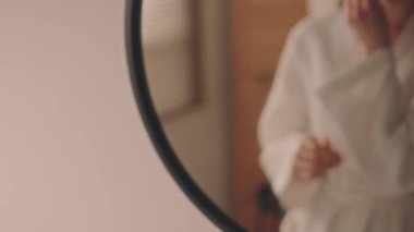 Beyaz cüppeli genç beyaz bir kadının sabah yüz masajı yaparken banyo aynasının önünde kuvars gua sha ile tenini düzeltirken, nefes açıcı rutinini düzeltirken.