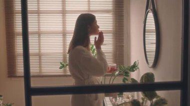 Sabahları bornozlu genç bir bayanın modern banyodaki aynanın önünde biyofilik tasarımla yüz kremi sürerken orta boy görüntüsü.