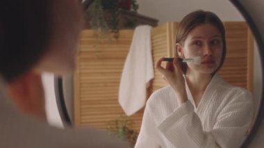 Kafkasyalı genç bir kızın banyo aynasının önüne silikon fırçayla yüz maskesi taktığı omuz aynası yansıması fotoğrafı, spa günü evde.