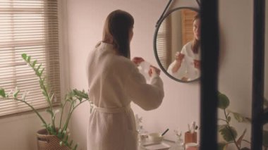 Bornozlu genç bir bayanın duş, cilt bakımı ve banyo aynası önünde yüz maskesi takması.