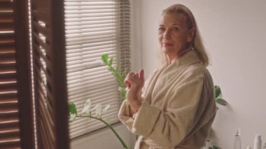 Banyodaki kameraya mutlu bir şekilde bakan, 60 'lı yaşlarda, bornozlu ve saç bantlı beyaz bir kadının orta boy portresi.