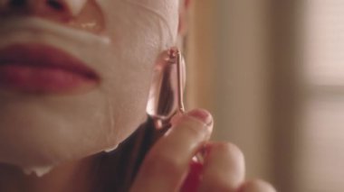 Yüz maskeli genç bayanın daha iyi soğurma, çene ve yanak masajı, cilt bakımı ve güzellik tedavisi için kuvars taşı silindiriyle masaj yaptığı son derece yakın plan.