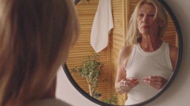 60 'lı yaşlarda emekli bir kadının kolsuz tişörtlü, taş silindirli yüz masajı yaparken, kırışıklık önleyici ve yaşlanmayı önleyici şeyler yaparken omuz aynası yansıması, güzellik tedavisi.