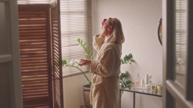 Modern banyoda pencereden bakarken yaşlanma karşıtı yüz kremi sürerken orta boy beyaz kadın pozu, 60 artı cilt rutini.
