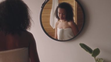 Havlu giymiş, aynadaki yansımaya bakan, duştan sonra terleme önleyici sprey sıkan, işe ya da okula, vücut bakımına ve güzelliğe hazırlanan iki ırklı genç bayanın arka görünüşü.