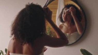 Havlulu kıvırcık saçlı genç bayanın arka görünüşü aynaya bakıyor ve duştan sonra silikon fırçayla yüzünü temizliyor. Banyoda, cilt bakımında ve güzellik tedavisinde.