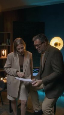 Kafkas erkek iş dünyasının podcast sunucusu ve kadın editör ya da yapımcısının stüdyoda yan yana durarak içerik planına baktığı ve kapsama için konuları tartıştığı orta boy dikey çekim.