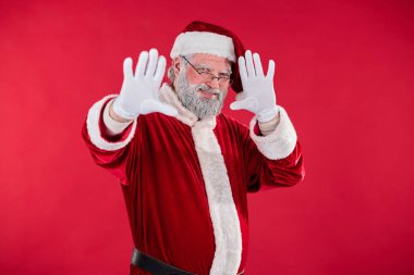 Noel Baba kılığında yaşlı bir adam gülümseyip el sallarken kırmızı arka planda neşeli bir ortam yaratıyor.