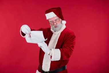 Noel Baba elinde kağıt ve kalem tutarken neşeli bir şekilde bir dilek listesi okuyor, kırmızı-beyaz geleneksel takım elbise giyiyor, canlı kırmızı arka plan önünde, Noel için hazırlanıyor.