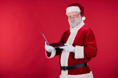 Kırmızı takım elbiseli ve şapkalı Noel Baba, gülümserken dizüstü bilgisayar tutuyor ve kullanıyor. Noel Baba neşeli bir şekilde teknolojiyle bağlantı kuruyor.