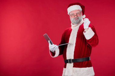 Kırmızı elbise giymiş Noel Baba kırmızı arka planda tablet tutuyor, kameraya gülümsüyor, şenlik ruhunu somutlaştırıyor. Çekici bir görünüş ve cazibe sunar.