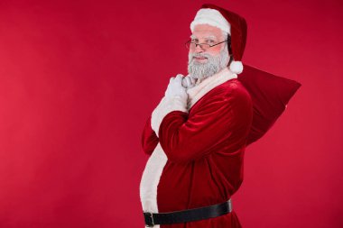 Noel Baba 'yı neşeli bir ifadeyle canlandırmak, klasik kırmızı ve beyaz giysiler içinde, kırmızı arka planda sakalını tutarken,