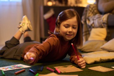 Genç kız rahat bir zeminde uzanıyor, rahat bir odada renkli kalemlerle çizimler yapıyor. Yastıklarla çevrili sanat eserleriyle meşgul ve yakındaki lambalardan hafifçe aydınlatılmış.