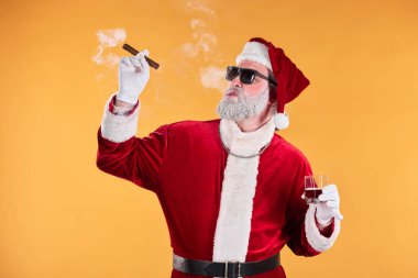 Yaşlı Noel Baba geleneksel kostümlü, elinde puro ve camla, sade sarı arka planda poz veriyor, neşeli ruhu ve rahatlığı somutlaştırıyor.