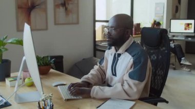 Yoğun, odaklanmış, günlük sweatshirt giyen, şirket ofisindeki masada çalışan, e-posta ya da program yazarken daktilo kullanan genç Afrikalı erkek yazılım mühendisinin orta kavisli görüntüsü.