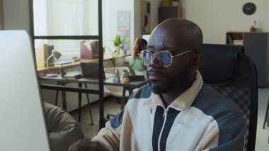 Orta ölçekli, rahat IT başlangıç ofisinde masa başında oturan, bilgisayarla uğraşan, sonra da gözlükleri ayarlayan, günlük eşofmanlı Afro-Amerikan bir adam.