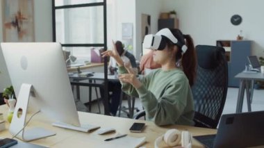 Asyalı kadın yazılım geliştiricisinin sanal gerçeklik kulaklığı taktığı, mobil uygulamaları test ederken boyutsal objeleri el hareketleriyle manipüle ettiği orta kavisli bir fotoğraf.
