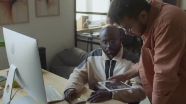 Orta boy Orta Doğulu erkek müteahhit bilgisayardaki kodları çalıştırırken masasının başında duruyor, sonuçlara bakıyor, ekrana işaret ediyor ve gelişmeleri tartışıyor.