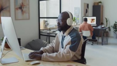 Engelli, tekerlekli sandalye kullanan, bilişim şirketinde masa başında oturan, yeni uygulamalar üzerinde çalışan, kameraya bakıp gülümseyen mutlu Afrikalı Amerikalı erkek yazılım geliştiricisinin orta boy portresi.