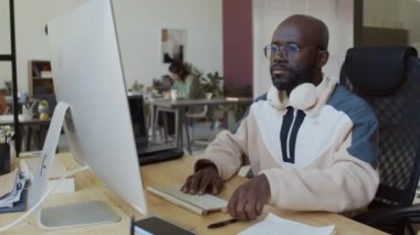 Gözlüklü bir Afrikalı Amerikalı programcının orta boy fotoğrafı, çalışma masasında oturan sweatshirt, masaüstü bilgisayarında yazılım kodunu test eden, algoritmalı deftere notlar yazan.