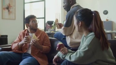 Üç etnik çeşitlilikte programcının yemek molasında kanepede oturup sandviç ve meyve yerken çekilmiş orta boy bir fotoğraf. Afro-Amerikalı erkek ve Çinli kadın, sürpriz meslektaşlarına haberleri veriyor.