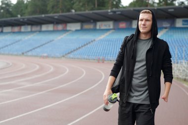 Spor şişesini elinde tutarken açık havada koşan atletik bir gencin portresi. Kendine güvenen ve yüzüne odaklanmış bir ifade.