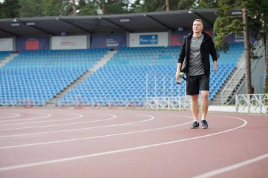 Spor kıyafetleri içinde, açık havada ısınan bir sporcu boş stadyumda antrenman yapmaya hazırlanıyor. Ağaçlar ve arka planda duran sakin atmosfer