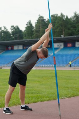 Atlet, ağaçlar ve stadyum koltuklarıyla çevrili atletizm pistinde sırıkla atlamaya hazırlanıyor. Odaklanmış ifade ve atletik duruş belirgin