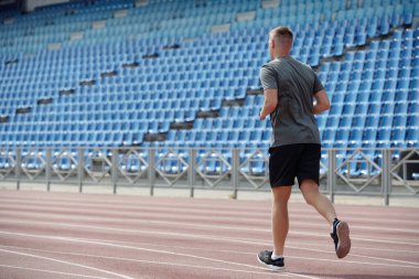 Spor giyim sektöründe sporcu adam mavi koltuklu boş stadyumda koşuyor, açık hava egzersizi ve fiziksel kondisyonuna odaklanıyor.
