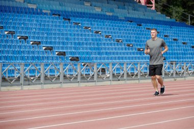 Gündüz vakti boş stadyumda koşan bir adam spor sahasında koşuyor ve arka planda mavi stadyum koltukları var.