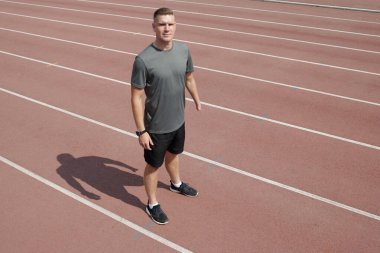 Athlete standing on track field wearing a gray t-shirt and black shorts. Preparing for athletic training outdoors. Shadow casted on running track clipart