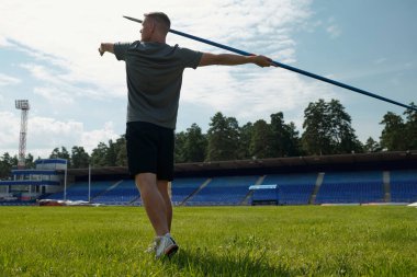 Yeşil ağaçlar ve açık mavi gökyüzü ile çevrili açık stadyumda spor kıyafetleri ve spor ayakkabıları giyerek cirit atan bir sporcu.
