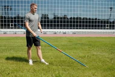 Spor sahasına cirit atmaya hazırlanan sporcu, günlük spor kıyafetleri ve spor ayakkabıları giyiyor, arka plandaki binalar cam panellere yansıyor.
