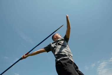 Çimenli bir alanda elinde mızrakla duran sporcu, açık gökyüzü altında dinamik bir duruşta yakalandı.