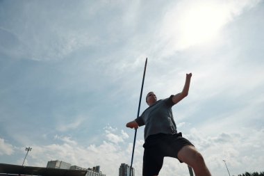 Arka planda şehir manzarası olan açık alanda sırıkla atlama çalışması yapan bir sporcu. Güneşli bir günde havadaki atletle kameraya bakmadan yapılan eylem