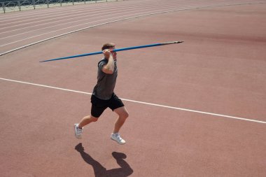 Athlete running on red track field holding javelin above shoulder and preparing release. Background includes part of stadium and fencing surrounding track field clipart