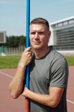 Arka planda modern bir binayla bir direğe tutunan bir adamın portresi, yeşil ve kırmızı renkli spor ortamını vurgulayan açık hava atletizm ortamını yakalıyor.