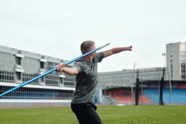 Athlete practicing javelin throw in sports complex, focusing on technique and form in preparation for competition. Background features grandstands and track clipart