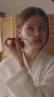 Kafkasyalı genç bir kızın banyo aynasının önüne silikon bir maskeyle yüz maskesi taktığı dikey bir fotoğraf, spa günü evde.