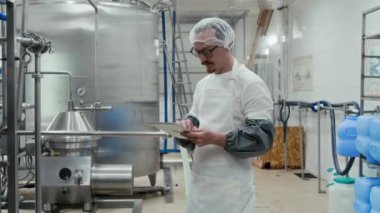 Orta boy beyaz üniformalı beyaz erkek peynir üreticisinin dijital tabletten kontrol edilen büyük endüstriyel otomatik süt depolama fıçısının yanında duruşu.