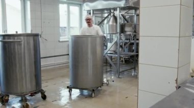 Beyaz önlüklü ve kapşonlu fabrika teknisyeni beyaz peynir üretirken uzun çelik süt arıtma fıçısını işleme tesisinde tekerlekler üzerinde taşırken tam tecrit görüntüsü.