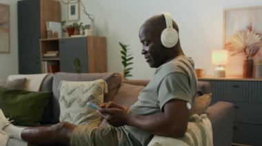 Orta yaşlı Afro-Amerikan bir adam kulaklık takıyor ve koluna sürekli glikoz monitörü takıyor. Evde kanepede dinlenirken müzik dinliyor ve akıllı telefon kullanıyor.