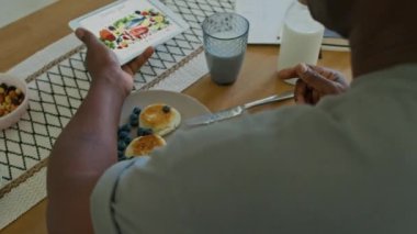 Kolunda CGM sensörü olan siyahi bir adamın dijital tableti diyabetli yiyeceklerle mutfak masasına yerleştirip kahvaltıda yaban mersinli kuark gözlemesi yediğini yakın plan.