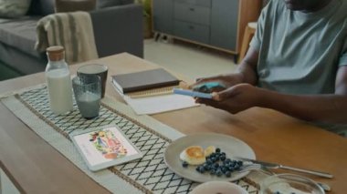 Afro-Amerikalı adamın akıllı telefondaki düşük glikoz seviyesini kontrol ederken yakın plan çekimleri, sonra da evde kahvaltı sırasında mutfak masasında otururken not alırken.