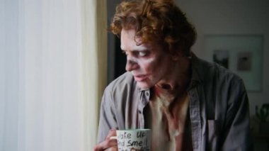 Kanlı, çürümüş vücutlu, korkunç beyaz gözlü, pencereye doğru yürüyen, dışarı bakan, iç çeken, koyu mizah desenli kahve içen, sonra da ağızlarını eliyle silen üzgün zombi adamın orta ölçekli yakın çekimi.