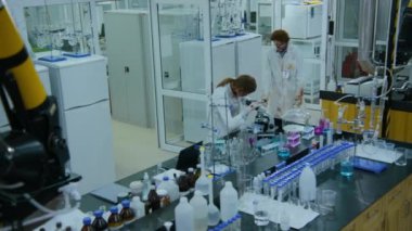 Yüksek açılı geniş açılı, kirli kanlı beyaz laboratuvar önlüğüyle yürüyen çürümüş et laboratuvara giriyor, kadın meslektaşının mikroskobu kötüye kullanmasına katılıyor ve dizüstü bilgisayarını yumrukluyor.